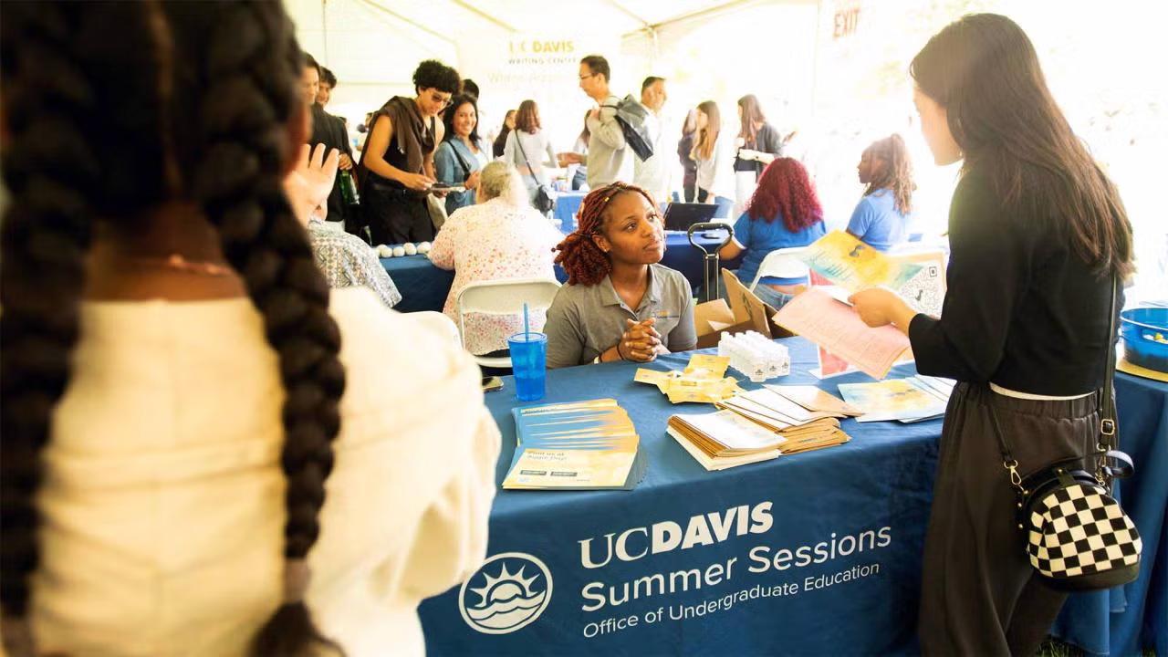 A lively informational fair under a large tent, where students engage with representatives at various tables. In the foreground, a woman behind a UC Davis Summer Sessions table speaks with a student holding informational flyers. The table is covered with brochures, flyers, and giveaways. Other tables and attendees are visible in the background, contributing to a busy and energetic atmosphere. The UC Davis Summer Sessions banner includes the logo and reads 'Office of Undergraduate Education.