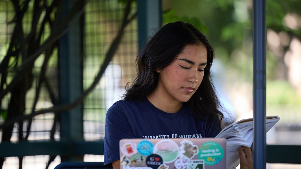 A student seated at an outdoor table with her laptop in front of her while she looks through a notebook.