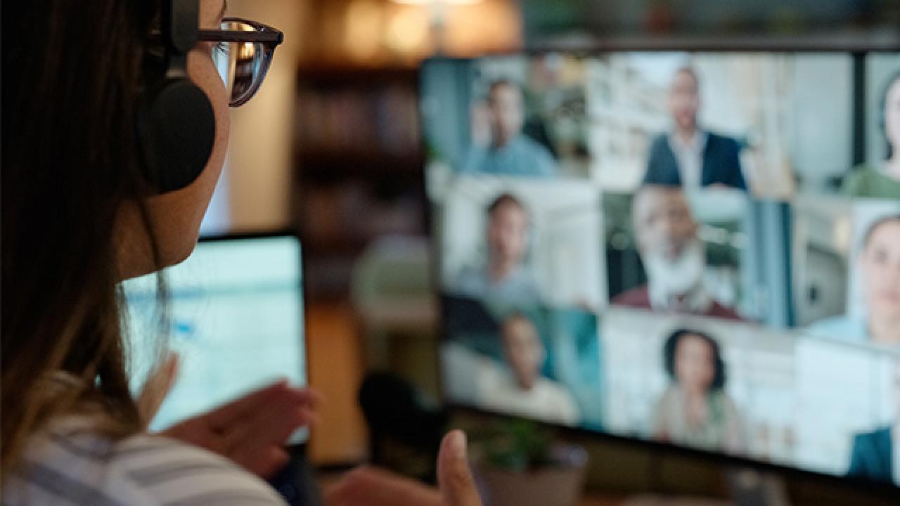 A person wearing a headset and glasses participates in a virtual meeting, gesturing while looking at a computer screen displaying multiple participants in a video call.