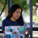 A student seated at an outdoor table with her laptop in front of her while she looks through a notebook.