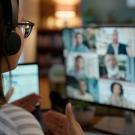 A person wearing a headset and glasses participates in a virtual meeting, gesturing while looking at a computer screen displaying multiple participants in a video call.